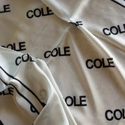 A close-up of a white or cream satin-like fabric with an allover black 'COLE' logo print and black border lines, draped with a hand visible at the bottom edge.