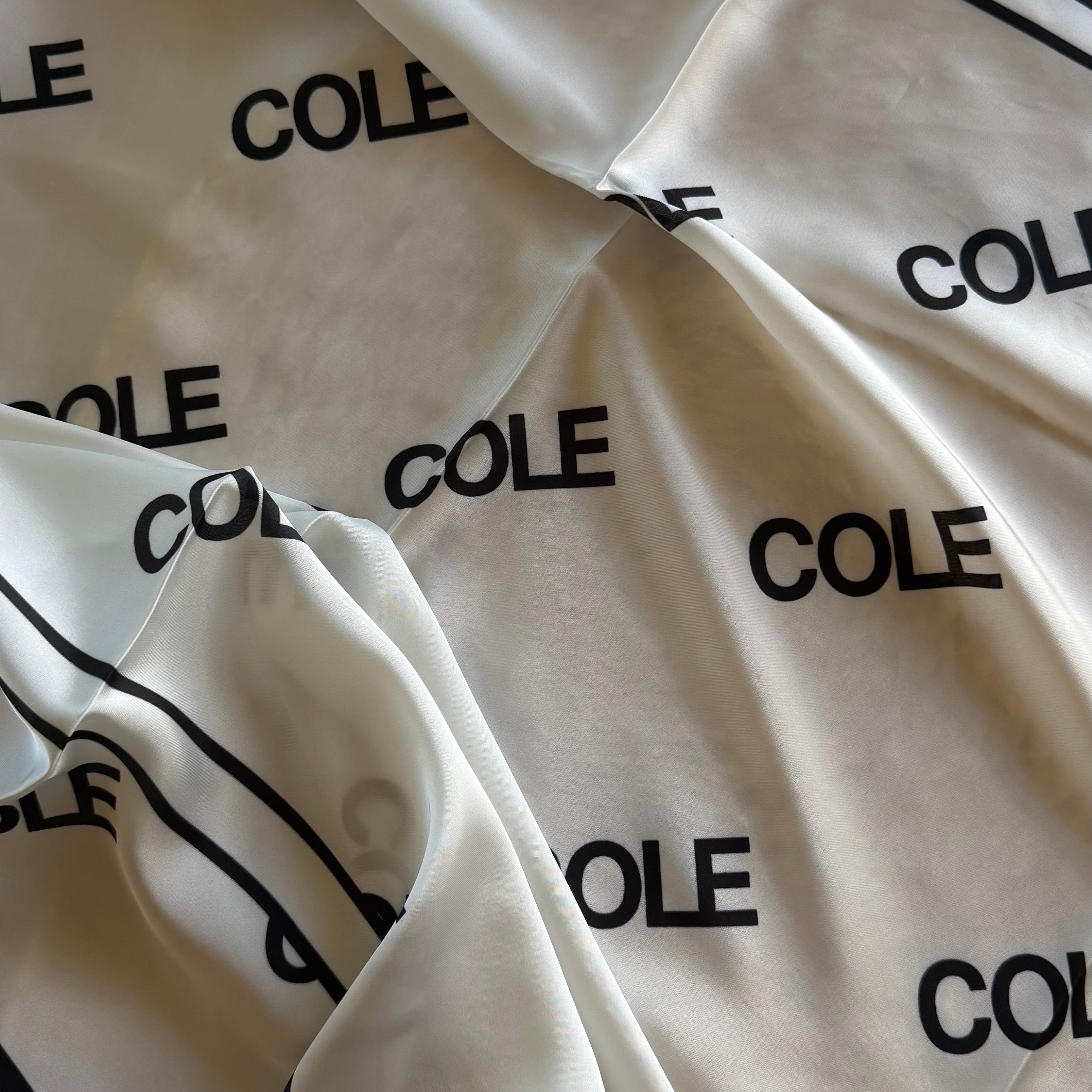 A close-up of a white or cream satin-like fabric with an allover black 'COLE' logo print and black border lines, draped with a hand visible at the bottom edge.